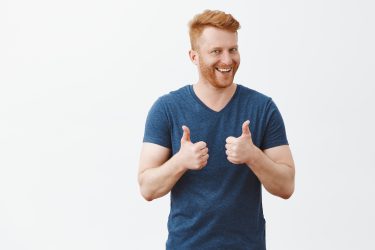 Nice job man, like it. Pleased good-looking happy male friend with ginger hair and brislte, showing thumbs up and smiling broadly, giving positive feedback, sharing his positive opinion over grey wall.